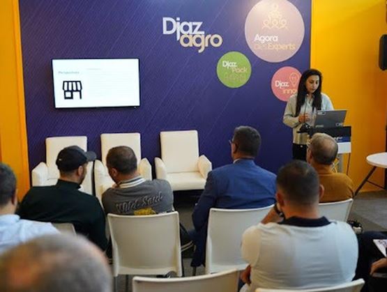 Une intervenante présente une conférence sur un stand aux couleurs de « Djazagro », face à un public assis, avec un écran affichant une diapositive et des logos tels que « Agora des Experts » et « Djaza Pack ».