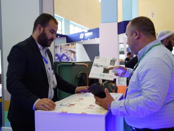 Deux professionnels échangent sur un stand d’exposition lors d’un salon, consultant des documents techniques sur un comptoir, avec des équipements présentés en arrière-plan.
