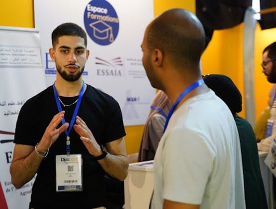 Deux participants échangent sur un stand « Espace Formation » lors d’un salon professionnel, portant des badges autour du cou, avec des panneaux d’information et des logos visibles en arrière-plan.