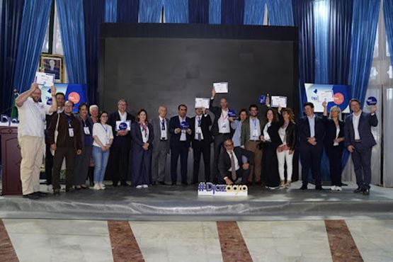 Un groupe de professionnels pose sur une scène lors d’un salon, certains tenant des documents ou des trophées, devant un grand écran et des rideaux bleus, avec le hashtag « #Djazagro » affiché au sol.