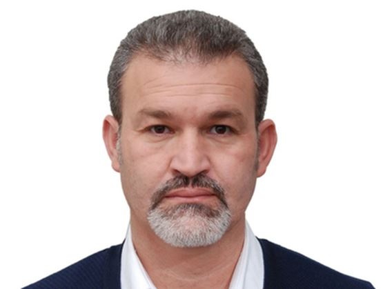 Portrait d’un homme avec des cheveux courts grisonnants et une barbe taillée, portant une chemise blanche et un cardigan sombre, regardant l’objectif avec une expression neutre sur fond clair.