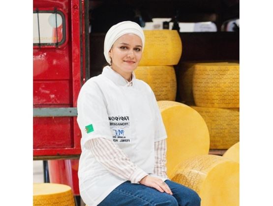 Une femme portant une tenue blanche et un couvre-chef est assise devant de grandes meules de fromage, dans un environnement de production ou d’exposition, avec un véhicule rouge en arrière-plan.