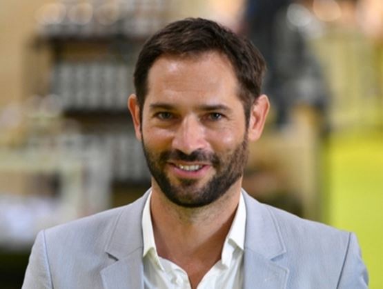 Portrait d’un homme souriant avec une barbe courte, portant une chemise blanche et une veste claire, sur un fond intérieur flou.