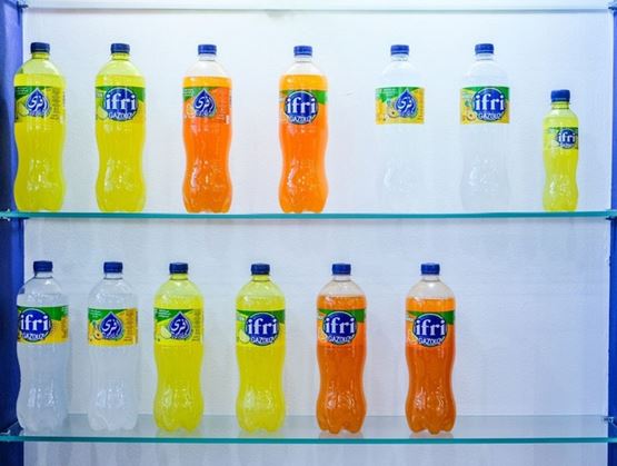 Bouteilles de boissons gazeuses Ifri Gazouz de différentes saveurs, alignées sur des étagères en verre dans un stand d’exposition.