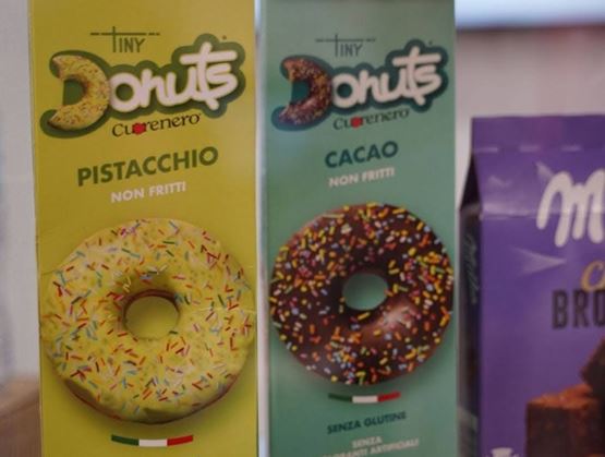 Boîtes de donuts emballés de la marque Tiny Donuts Cuorenero, saveurs pistache et cacao, présentées sur un stand, avec mention sans gluten et non frits.