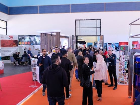 Allée d’un salon professionnel avec plusieurs visiteurs circulant entre des stands d’exposition, sur un sol recouvert de tapis rouge et orange, dans un hall lumineux.