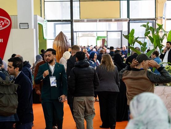 Allée animée d’un salon professionnel avec de nombreux visiteurs, des stands d’exposition et un sol orange.