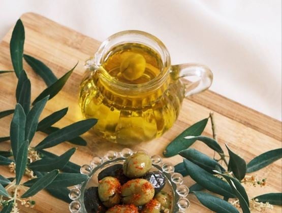 Pichet en verre rempli d’huile d’olive, accompagné d’olives vertes et noires et de rameaux d’olivier sur une planche en bois.