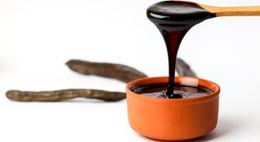 Carob molasses being poured into a terracotta bowl, with pods in the background.