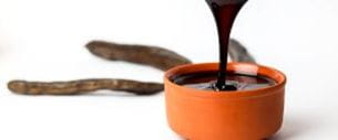 Carob molasses being poured into a terracotta bowl, with pods in the background.