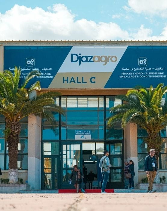 Entrance of the Djazagro trade show with a sign displaying "Hall C", surrounded by palm trees and visitors entering and exiting the building.