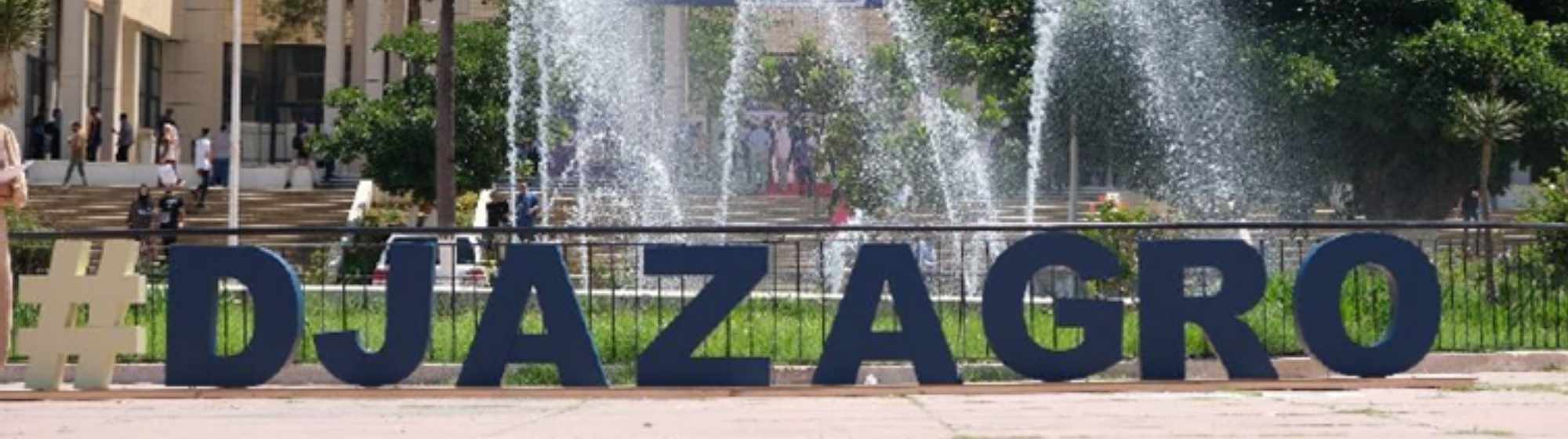 Lettres géantes #DJAZAGRO installées devant la fontaine du salon Djazagro, avec le bâtiment principal et les visiteurs en arrière-plan.